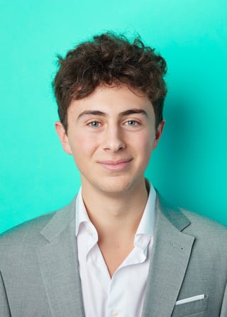 Young man with dark curly hair wearing gray blazer and white shirt against turquoise background in professional headshot