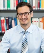 Professional portrait of a man wearing glasses, light blue shirt and striped tie, smiling at camera with bookshelves in background