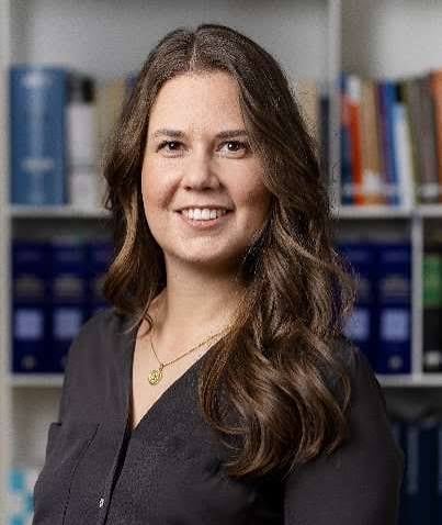 Professional headshot of a woman with long brown wavy hair wearing a dark cardigan against a bookshelf background